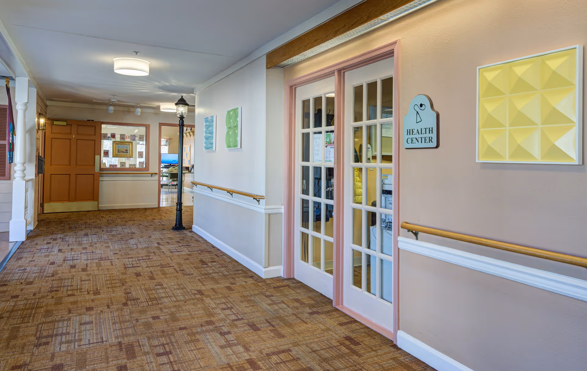 Interior hallway of Arden Courts - ProMedica Memory Care Community with carpeted floor, handrails on both sides, a lamp post style light fixture, and double glass doors labeled Health Center. The walls are decorated with colorful geometric art pieces.