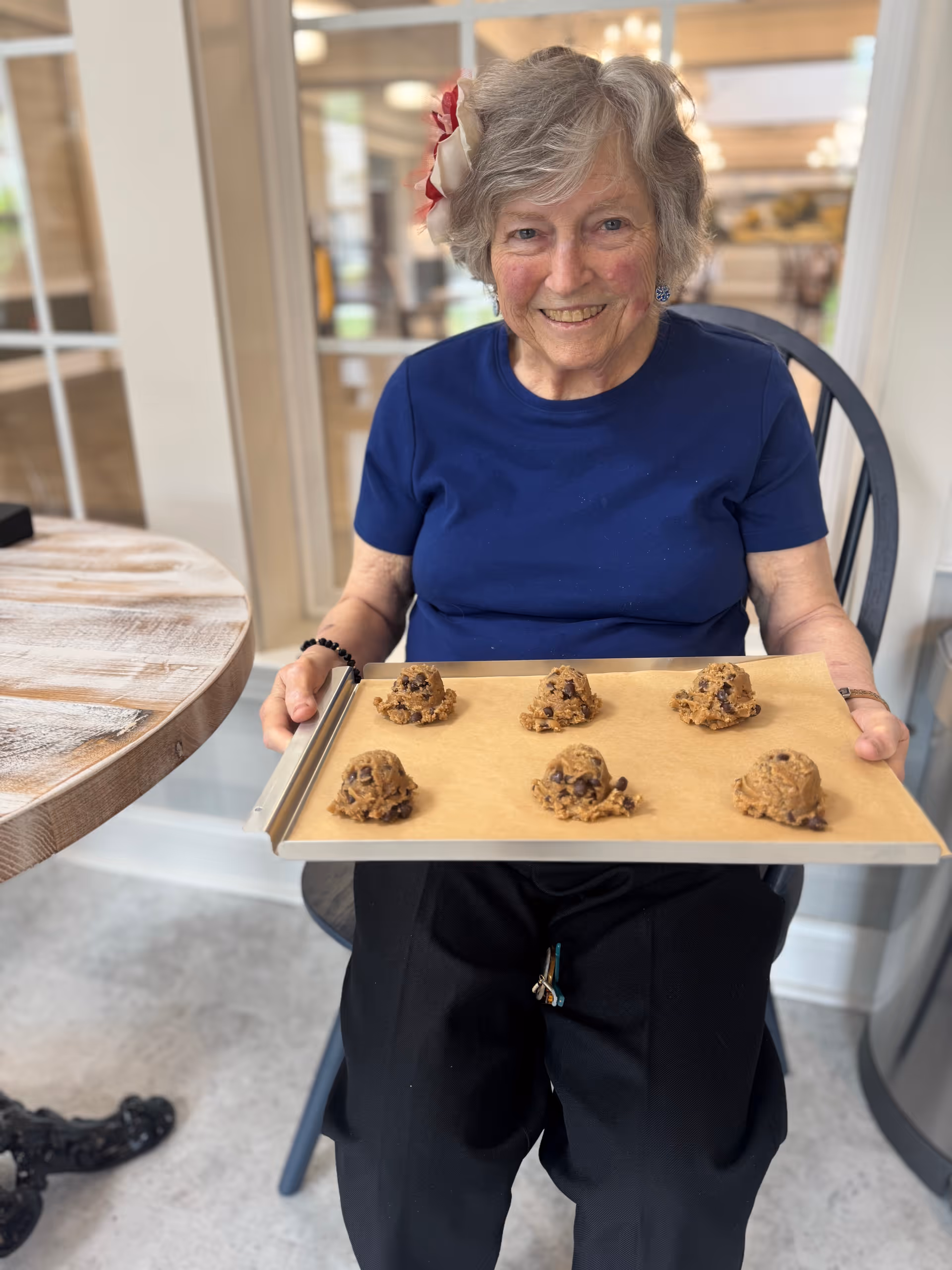 An elderly woman with gray hair and a flower hair accessory is sitting on a chair indoors, smiling and holding a baking tray with six scoops of chocolate chip cookie dough on parchment paper. She is wearing a blue shirt and black pants. The background shows a bright room with large windows and a wooden table nearby.