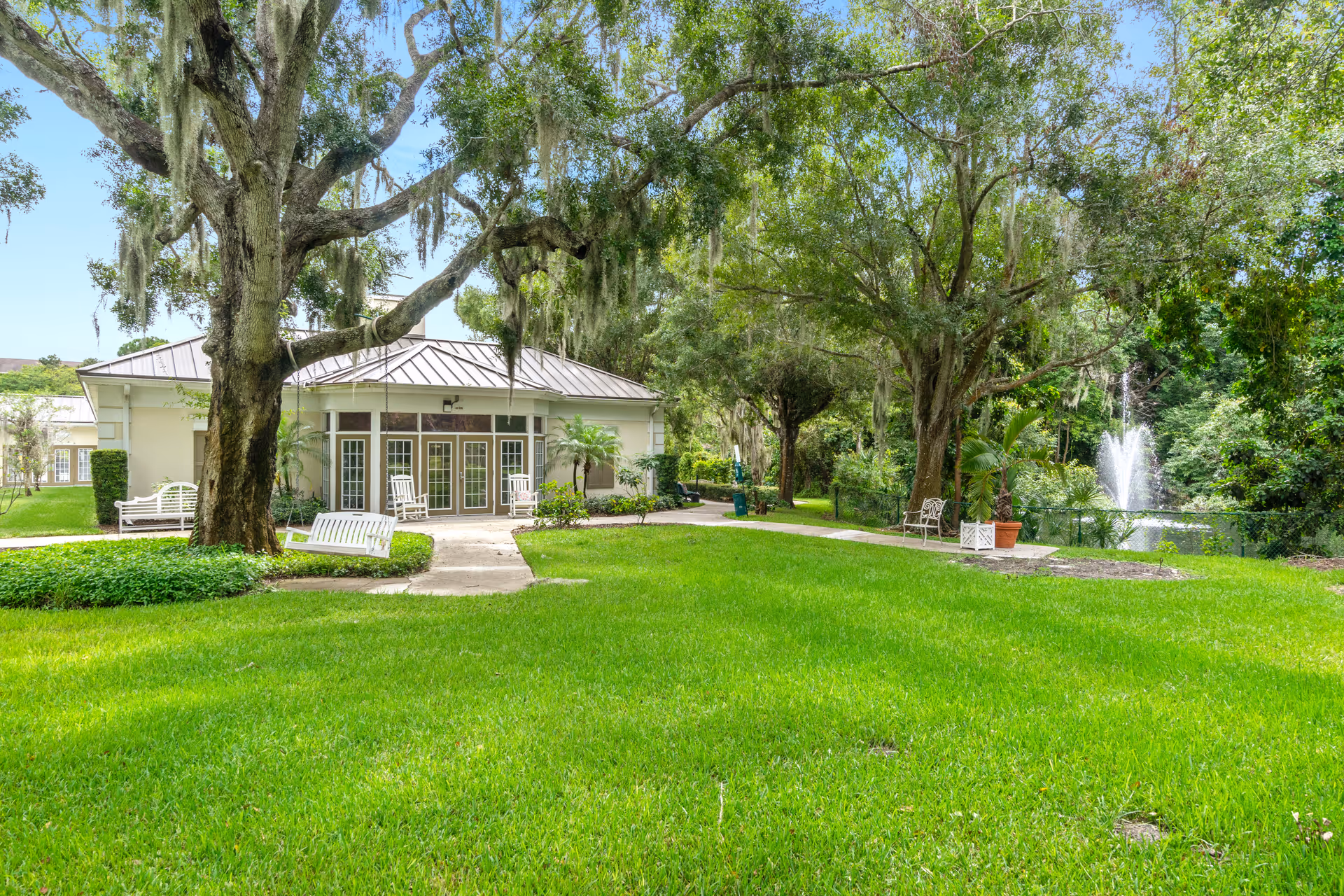 A lush green lawn with large trees and benches surrounding a building with multiple glass doors. There is a water fountain in a pond to the right, and the area is shaded by tall trees with hanging moss.