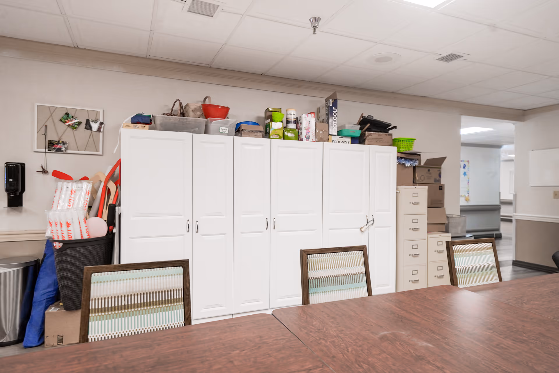 Long wooden table and chairs in front of white storage cabinets and stacked boxes in a communal activity/dining room.