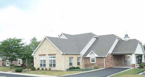 Exterior view of a single-story senior living facility building with a combination of stone and brick facade, multiple windows, a gray shingled roof, and a covered entrance. The building is surrounded by a paved driveway and some landscaping with small bushes and a tree.