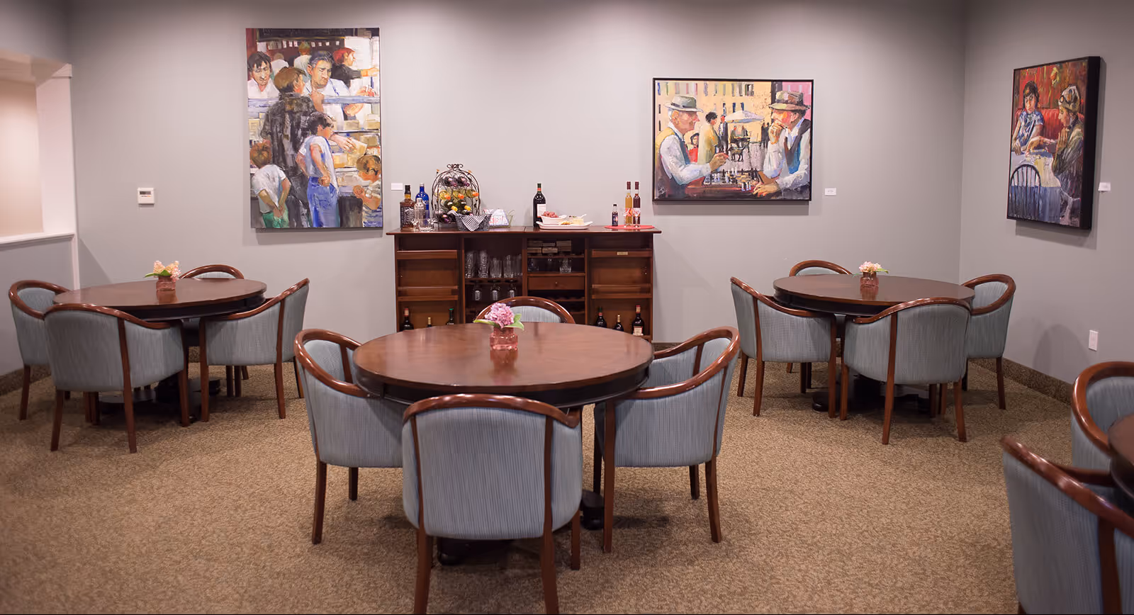 A dining room with three round wooden tables, each surrounded by six upholstered chairs. Each table has a small flower arrangement in the center. Against the back wall is a wooden cabinet with bottles of wine and glasses on top and inside. The walls are decorated with three colorful paintings depicting people in various scenes.