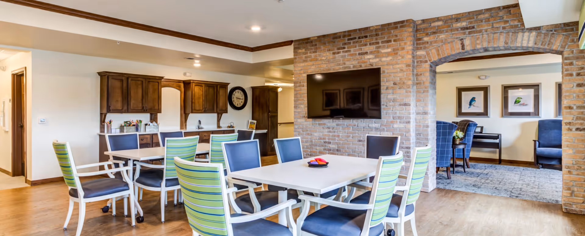 Communal dining area with multiple tables and green-striped chairs, a brick wall with a mounted TV, and a lounge visible through an archway.