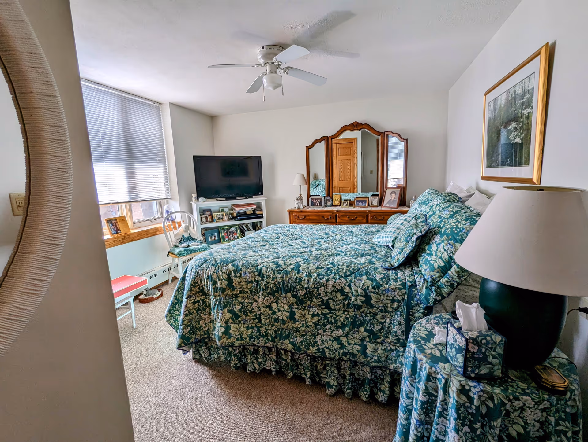 A cozy bedroom with a floral green bedspread and matching pillows. The room features a wooden dresser with a large mirror, several framed photos, a TV on a white stand, a white chair with a cushion, and a round side table with a lamp and tissue box. A framed picture hangs on the wall, and a window with blinds lets in natural light.
