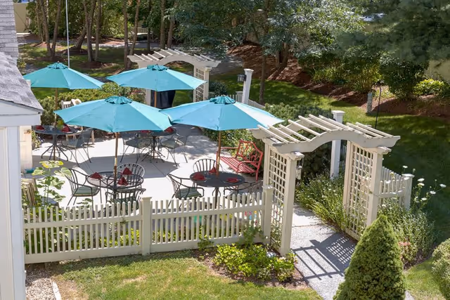 Outdoor patio area with several round tables and chairs under teal umbrellas, surrounded by a white picket fence and garden greenery, with a white wooden arbor entrance.