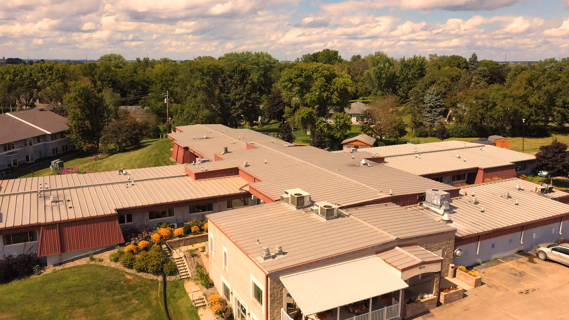 Aerial view of Green Prairie Rehabilitation facility showing a large single-story building with a flat roof surrounded by green lawns, trees, and a parking area with a few cars. The sky is partly cloudy.