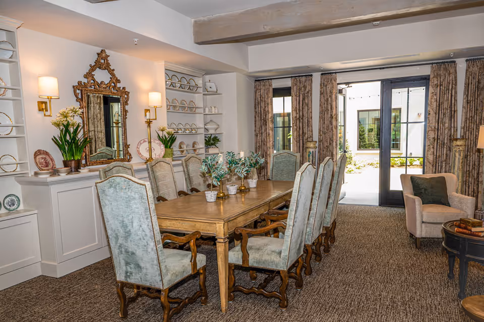 A well-lit dining room with a long wooden table surrounded by eight upholstered chairs. The room features built-in shelves displaying decorative plates and vases, a large ornate mirror on the wall, and two wall-mounted lamps. There are patterned curtains framing glass doors that lead to an outdoor area. A comfortable armchair and a small round table with books are visible in the corner.