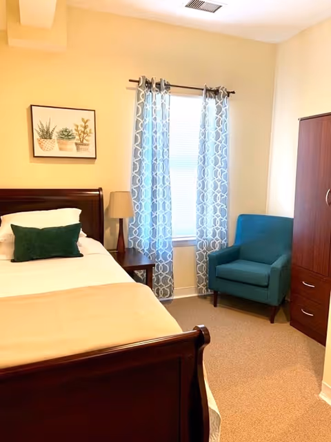 A cozy bedroom with a wooden bed frame, white bedding, and a green pillow. Next to the bed is a wooden nightstand with a lamp. A window with patterned curtains lets in natural light. There is a teal armchair in the corner and a wooden wardrobe with drawers on the right side of the room. A framed picture of potted plants hangs on the wall above the bed.