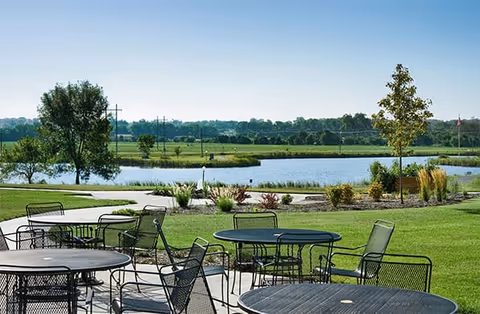 Outdoor patio area with several black metal tables and chairs on a concrete surface, overlooking a grassy lawn, a pond, and trees in the background under a clear blue sky.