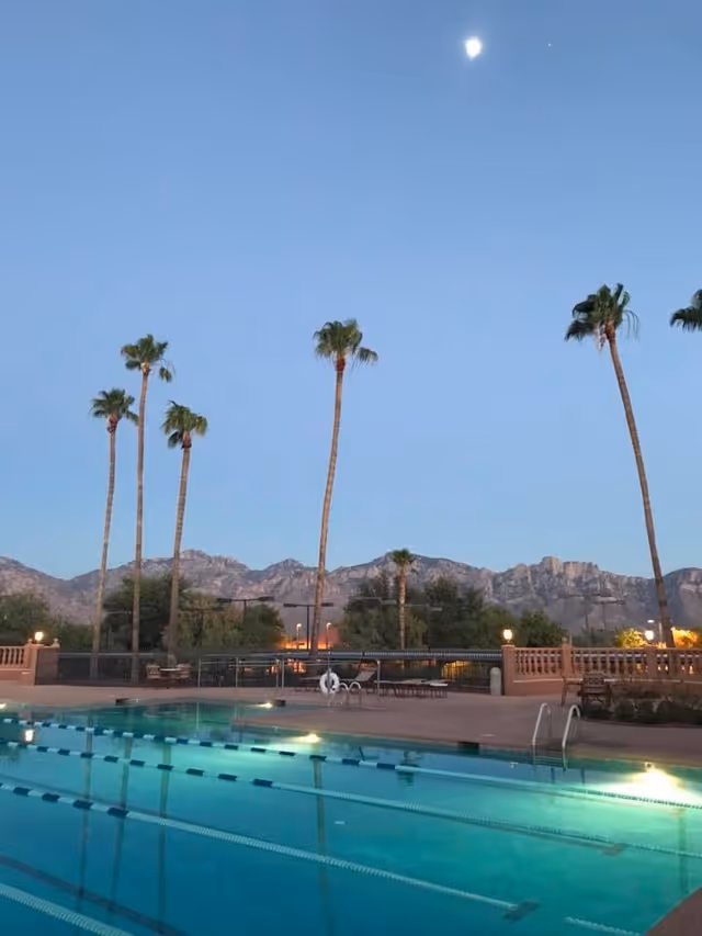 Outdoor swimming pool at dusk with tall palm trees, mountains in the background, and the moon in the sky.