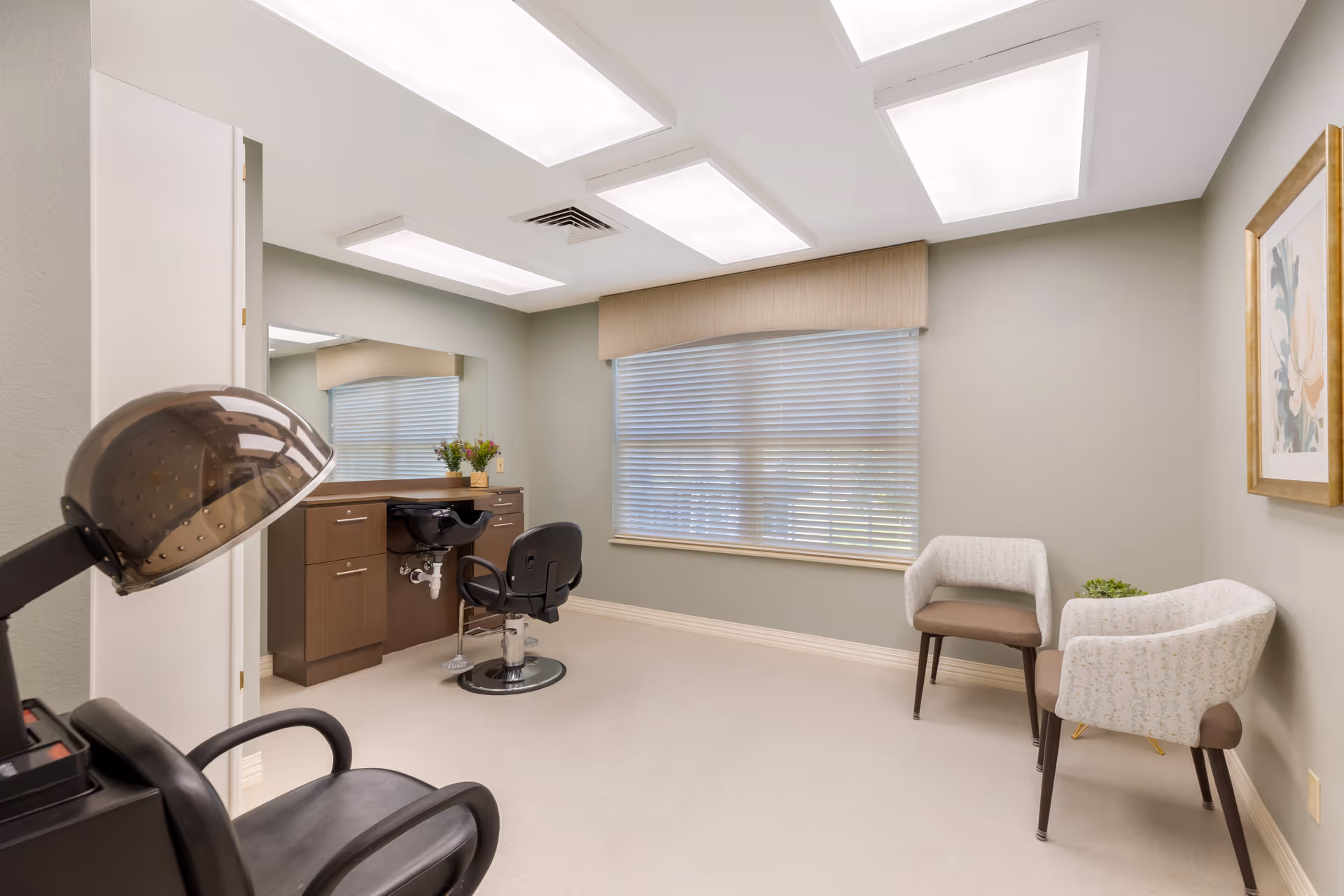 A bright salon-style interior with a styling chair, shampoo sink, hooded hair dryer, mirror and two waiting chairs by a window.