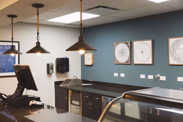 Interior view of a modern kitchen or food service area with dark countertops, three hanging pendant lights, a computer terminal, a hand sanitizer dispenser, and framed abstract artwork on a teal accent wall.