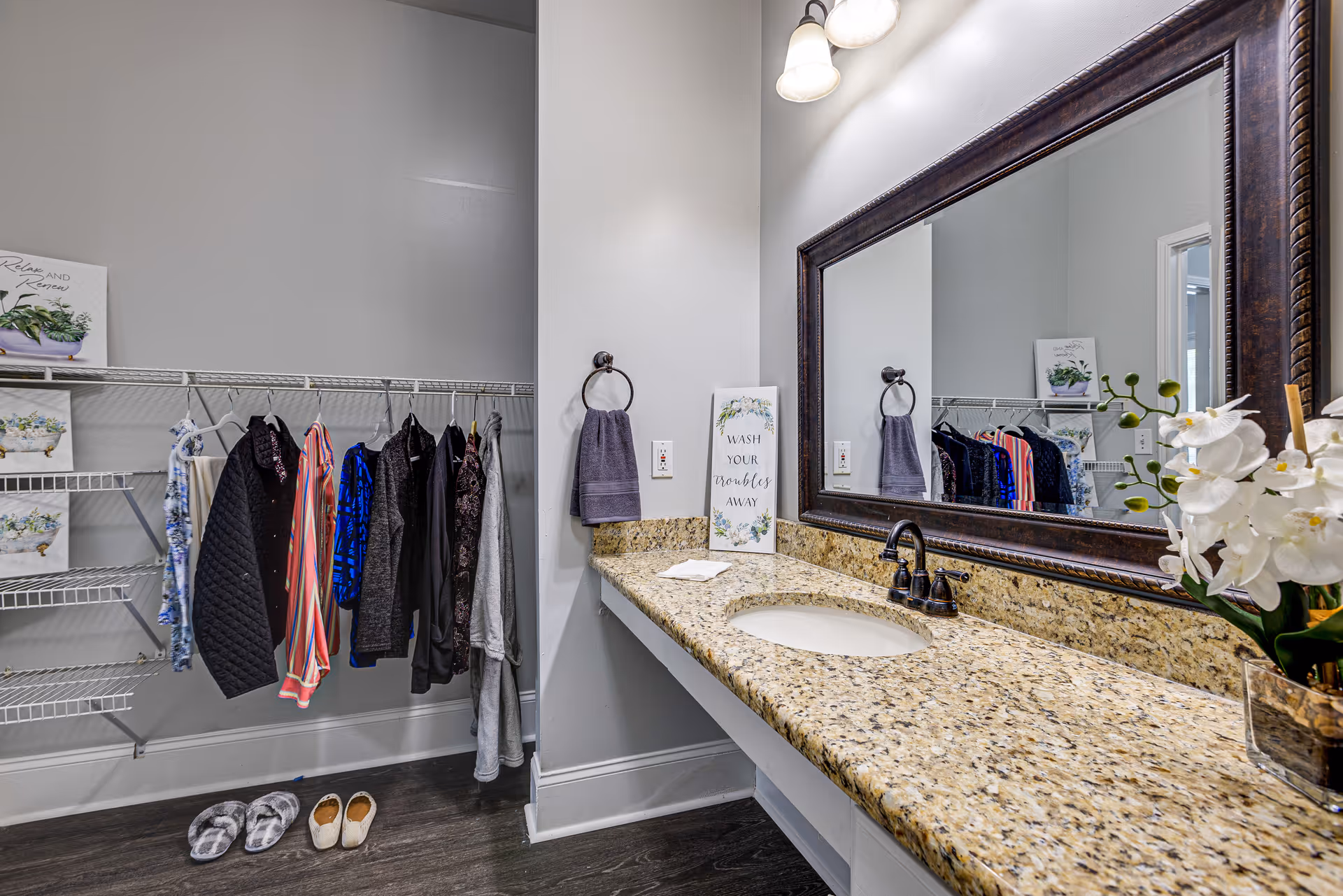 A bathroom vanity area with a granite countertop, an undermount sink, a large framed mirror, and a light fixture above. To the left, there is an open closet with hanging clothes and shelves, along with slippers on the floor. A decorative sign on the counter reads 'Wash your troubles away,' and a purple hand towel hangs on a ring beside the sink. A vase with white flowers is placed on the right side of the countertop.