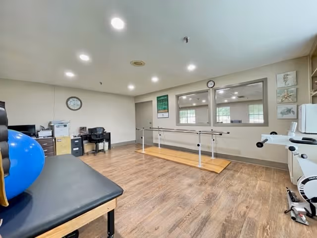 A rehabilitation room with wooden flooring, featuring a black padded therapy table with a blue exercise ball on it, parallel bars for walking exercises, an exercise bike, and a desk area with office chairs and equipment. The walls are light-colored with a clock and a calendar, and there are large windows looking into another room.