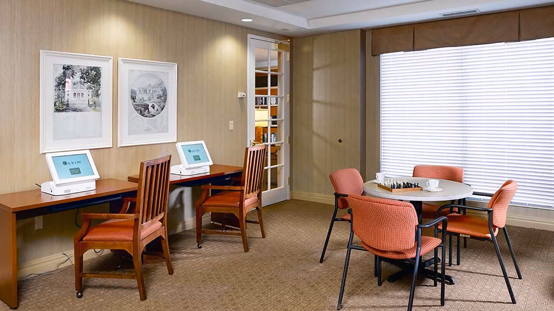 A bright communal room with two computer stations along a wall and a round table with four orange chairs and a chess set by a large window.