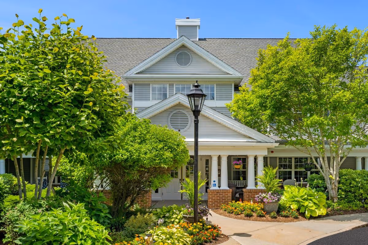 Front exterior view of a senior living facility named Sunrise of Plymouth Beach, featuring a well-maintained garden with various green trees and colorful flowers, a paved walkway leading to the entrance with white columns, and a black lamppost in the center.