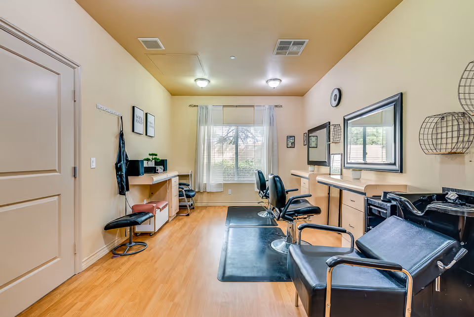 Interior view of a senior living facility's hair salon with wooden floors, beige walls, and a large window with sheer curtains. The room contains black salon chairs, mirrors mounted on the wall, a hair washing station, and various salon equipment and decor.