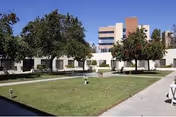 Outdoor courtyard area with green grass, paved walkways, and several trees. In the background, there is a multi-story building and a single-story building surrounding the courtyard.