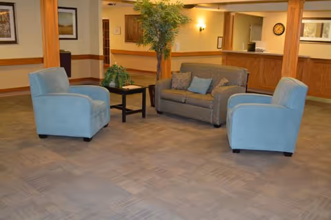 A seating area in a nursing and rehabilitation facility with two light blue armchairs and a patterned loveseat arranged around a small black coffee table. There is a tall artificial plant behind the loveseat, and the background shows a reception desk with a clock on the wall and framed artwork.