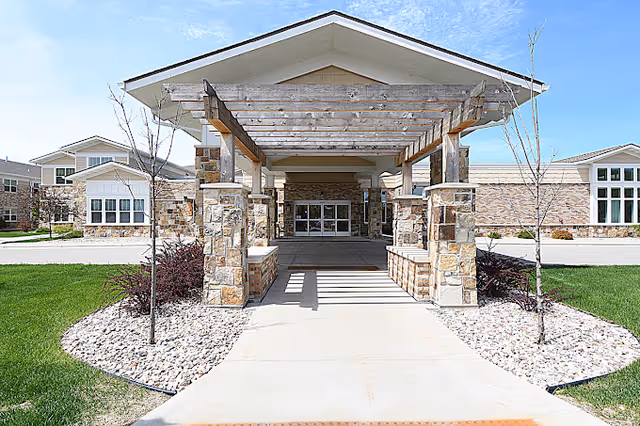 Front exterior view of Oak Park Place Menasha facility showing a covered entrance with stone pillars and a wooden pergola structure, surrounded by landscaped grass and small trees under a blue sky.