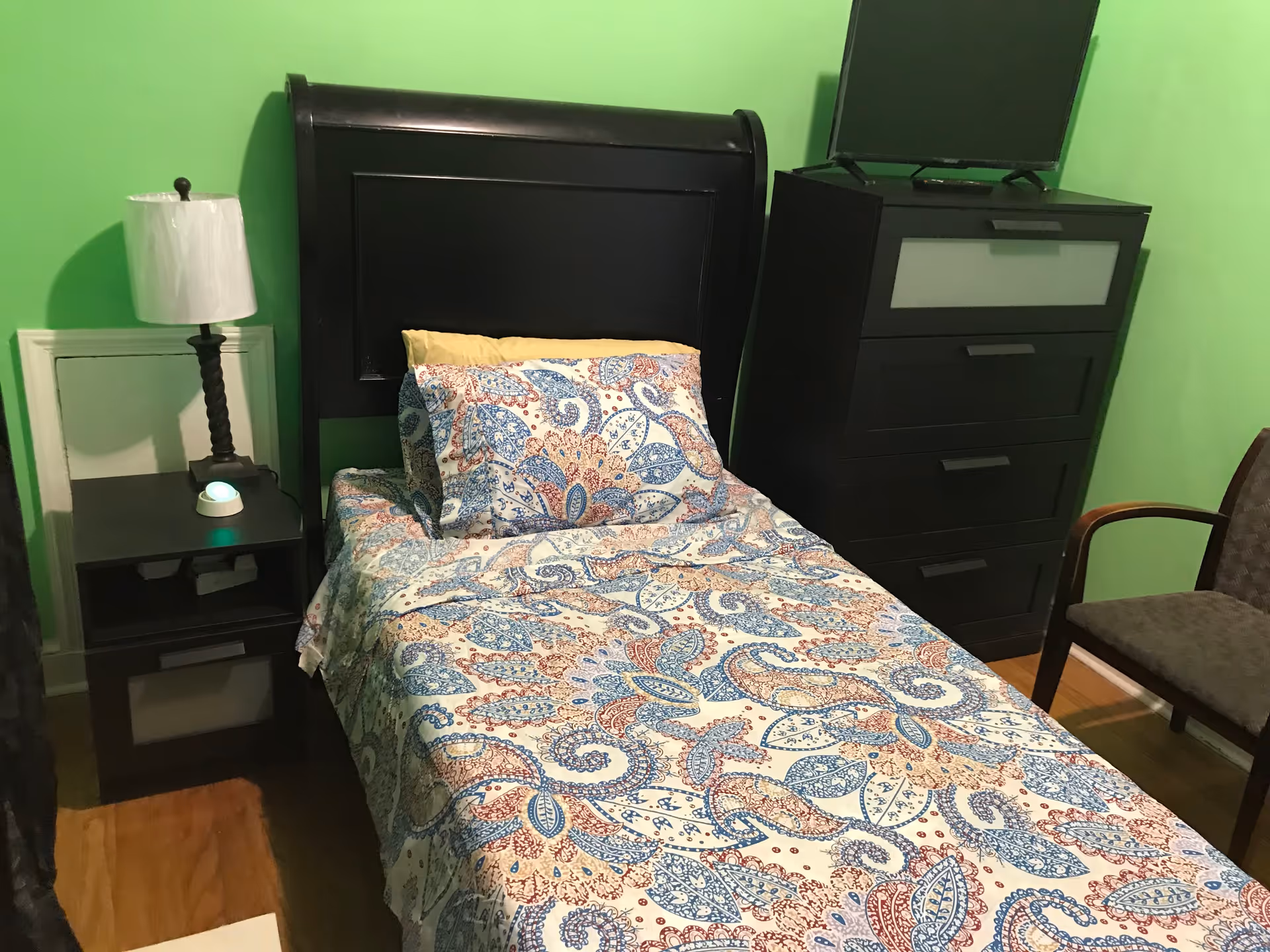 A small bedroom with a single bed featuring a black headboard and colorful paisley-patterned bedding in blue, red, and beige. Next to the bed is a black nightstand with a lamp and a small device with a green light. Across from the bed is a black dresser with a flat-screen TV on top. A wooden chair with a cushioned seat is positioned near the dresser. The walls are painted bright green and the floor is wooden.