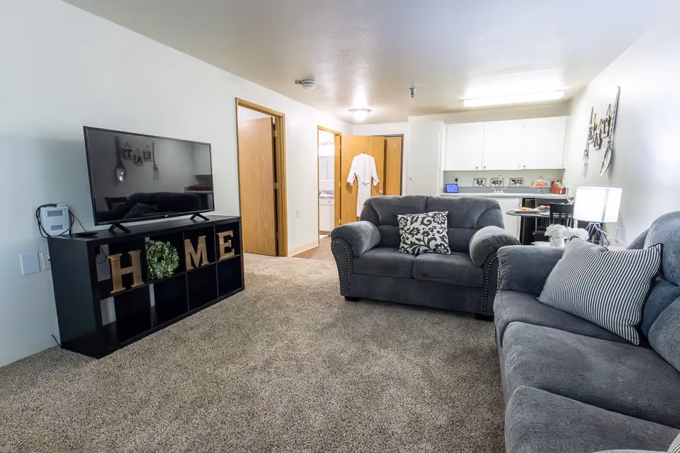 A cozy living room in an independent living facility featuring two gray upholstered sofas with patterned throw pillows, a black TV stand with decorative letters spelling HOME, a flat-screen TV, and a view into a small kitchen and hallway with open wooden doors.
