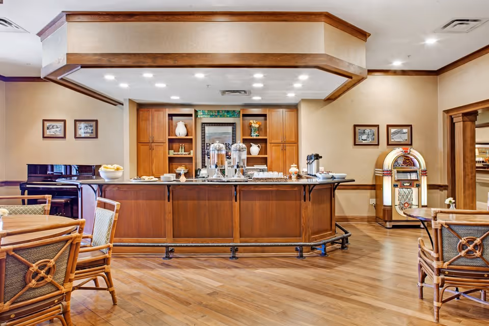 A cozy dining area in a senior living facility featuring a wooden buffet counter with beverage dispensers, cups, and plates. The room has wooden flooring, wicker chairs with cushions around tables, a vintage jukebox in the corner, framed pictures on the walls, and a piano in the background.