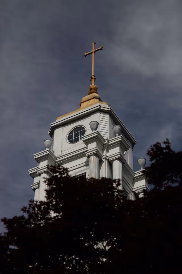 Close-up view of the top section of a white church steeple with classical columns and a round window, topped with a golden cross against a cloudy sky, partially obscured by dark tree branches.
