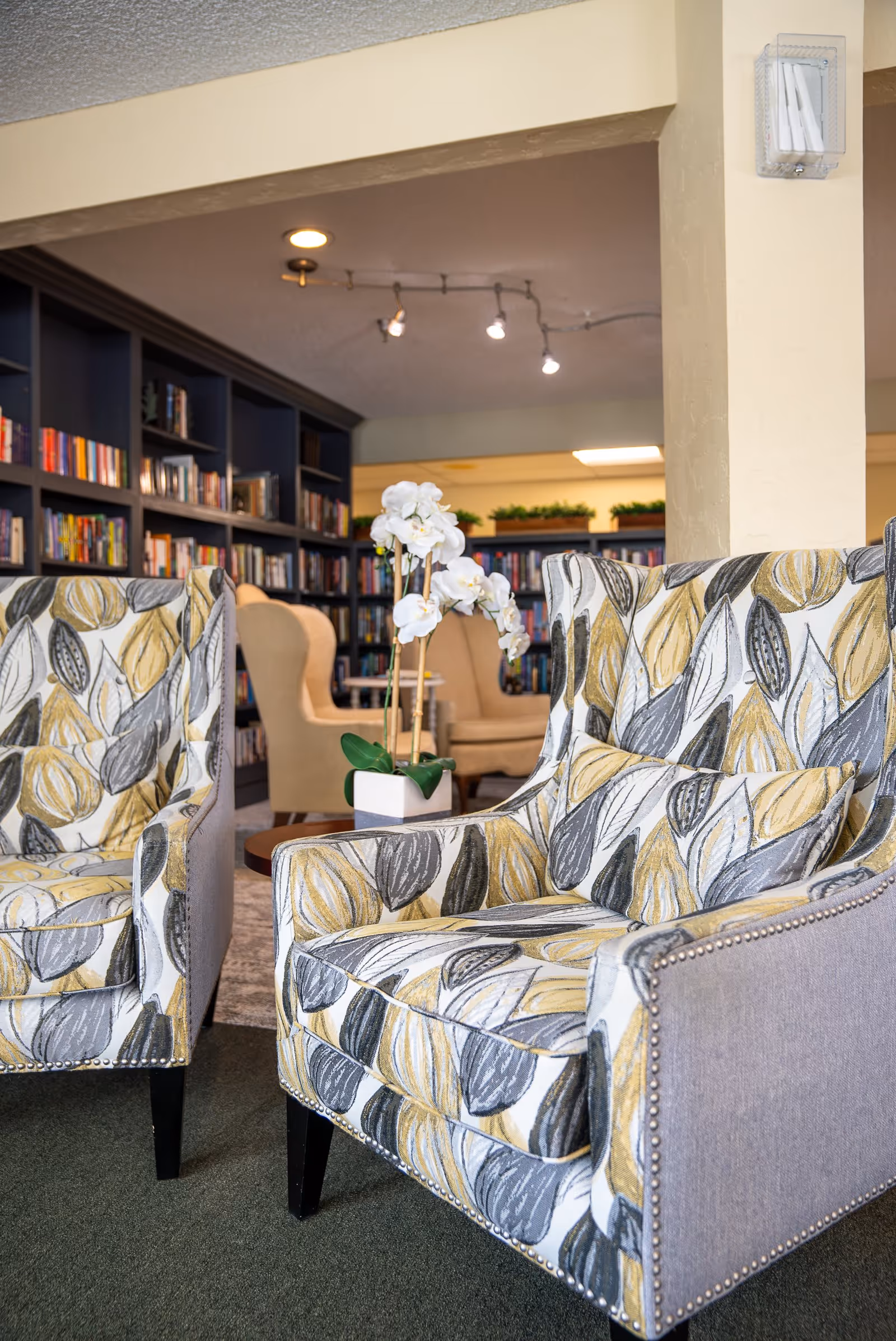 Two patterned armchairs with leaf designs in yellow, gray, and white colors are positioned in a cozy seating area. Behind them, there are beige wingback chairs and bookshelves filled with books, creating a warm and inviting library or lounge space.