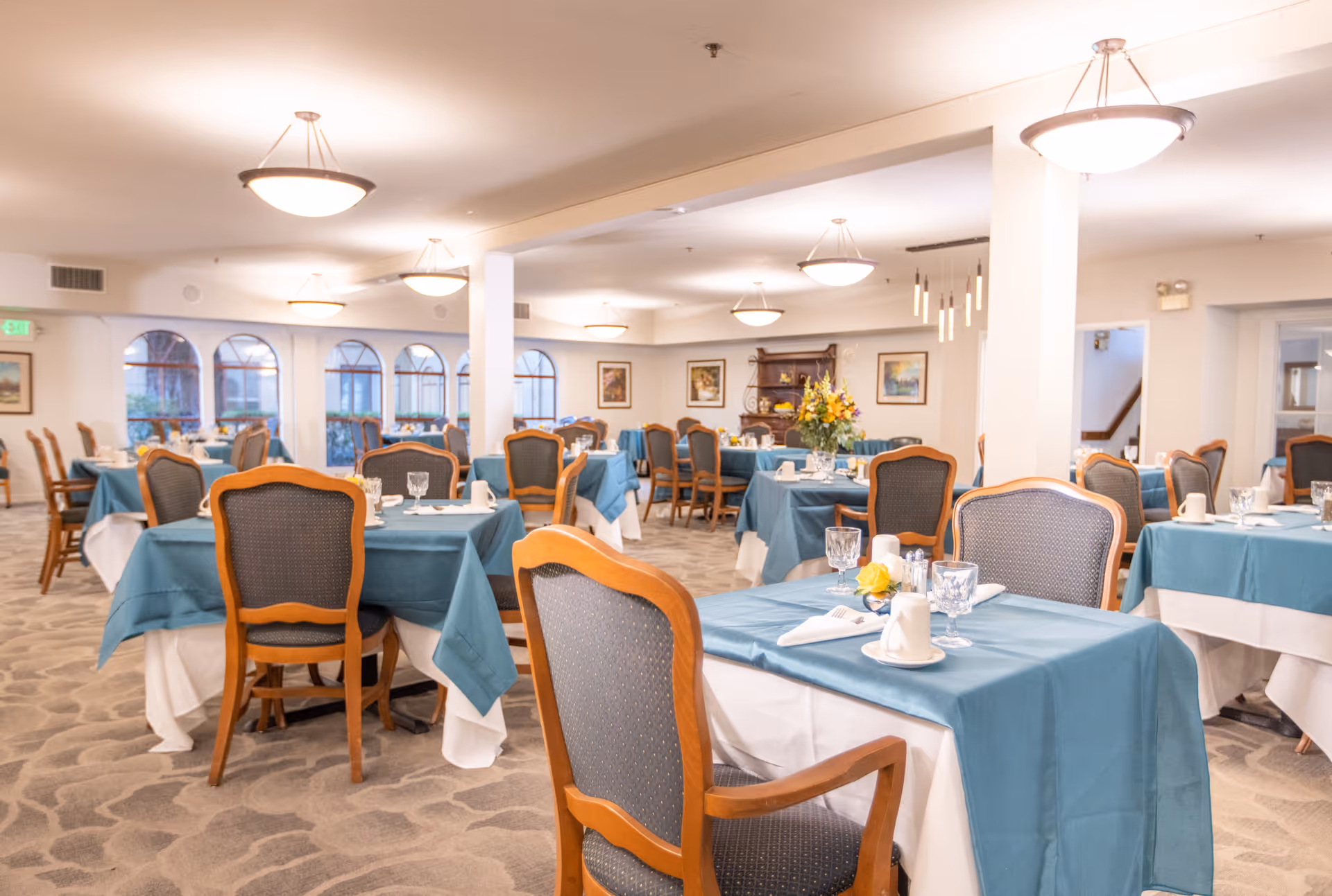A spacious dining room with multiple tables covered in teal tablecloths and white underlays, each set with glassware, napkins, and cups. The room features wooden chairs with cushioned seats and backs, soft lighting from ceiling fixtures, and large windows with arched frames letting in natural light. There are framed paintings on the walls and a floral centerpiece on one of the tables.