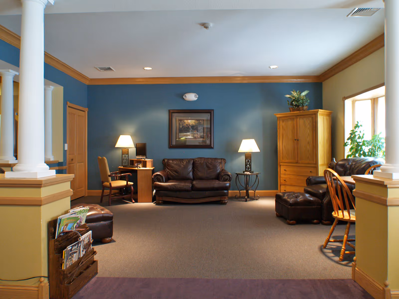 A cozy living room area with a brown leather sofa and matching armchair with ottoman, two table lamps on either side of the sofa, a wooden cabinet with a plant on top, a small desk with a chair and computer, and a magazine rack near white columns. The walls are painted blue and yellow with wood trim, and there is a framed picture hanging above the sofa.