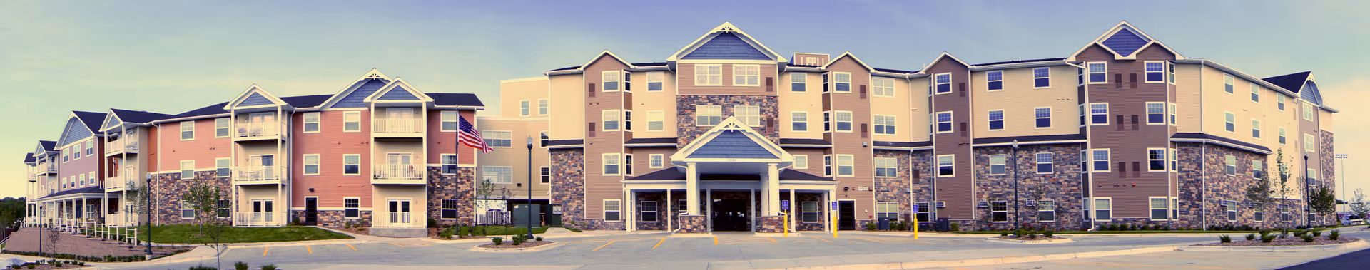 Wide panoramic view of Polar Ridge Senior Living building exterior featuring a multi-story structure with a combination of stone and siding facade, multiple balconies, and an American flag near the entrance.
