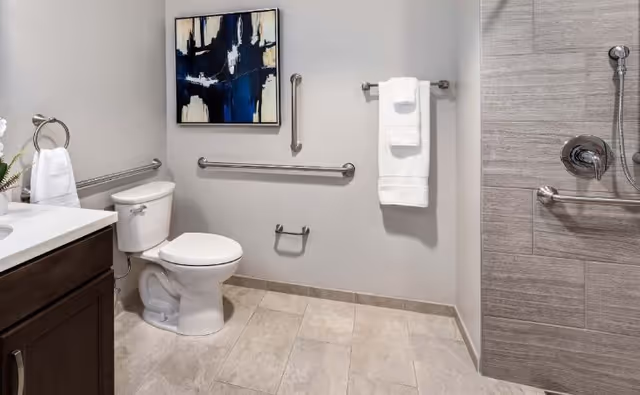 A clean and modern bathroom with a white toilet, a dark wood vanity with a white countertop, and a walk-in shower with gray tiles. The walls are light gray and feature stainless steel grab bars and towel racks with white towels. A blue and white abstract painting hangs above the toilet.