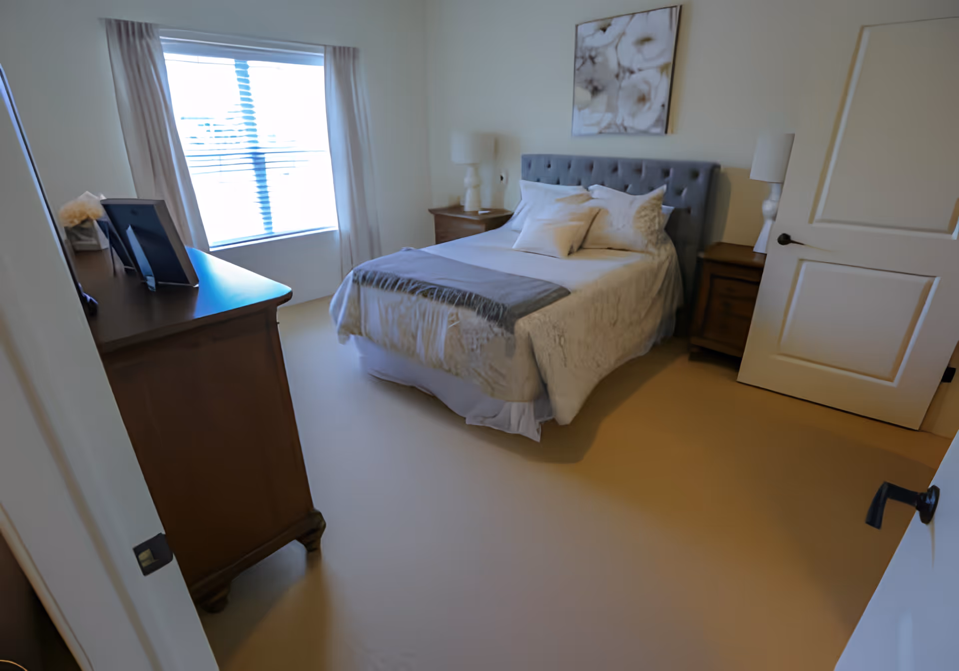 Sunlit bedroom with a neatly made bed, two nightstands with lamps, a dresser, and a window with curtains.