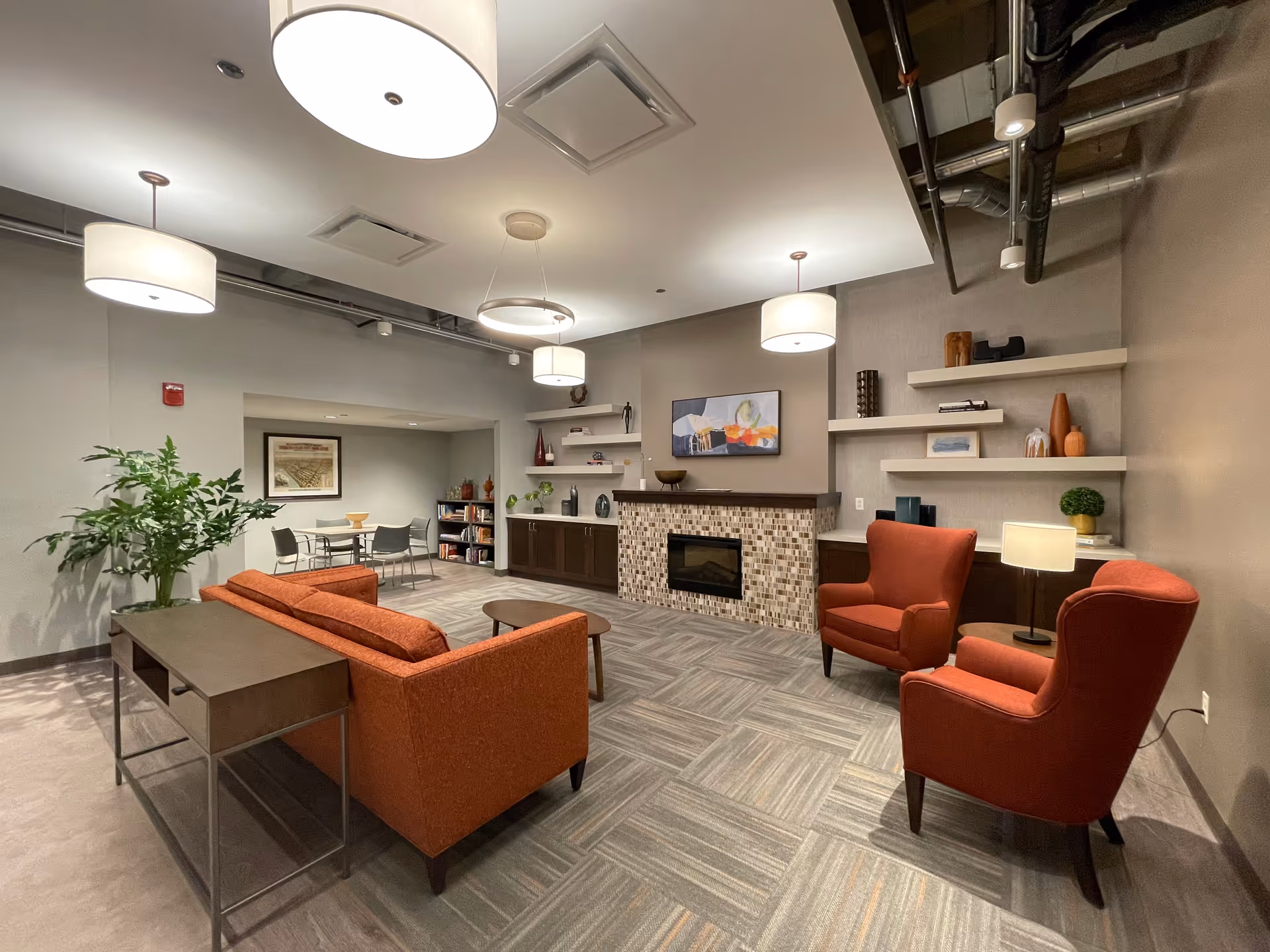 A modern and cozy living room area in a supportive living facility featuring an orange sofa, two matching armchairs, a small coffee table, and a fireplace with a mosaic tile surround. The room has built-in shelves with decorative items, a large potted plant, and a small dining table with chairs in the background. The ceiling has several round pendant lights and exposed ductwork.