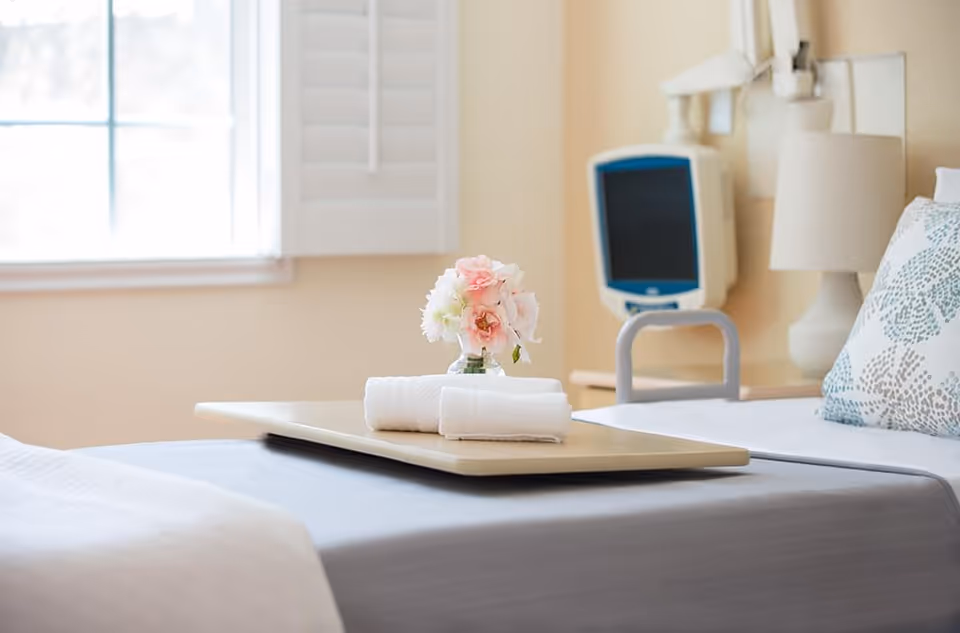 A neatly made hospital-style bed with a tray on top holding a small vase of pink and white flowers and two rolled white towels. The bed has a patterned pillow and a bedside lamp, with a medical monitor mounted on the wall behind it. A window with white shutters lets in natural light.