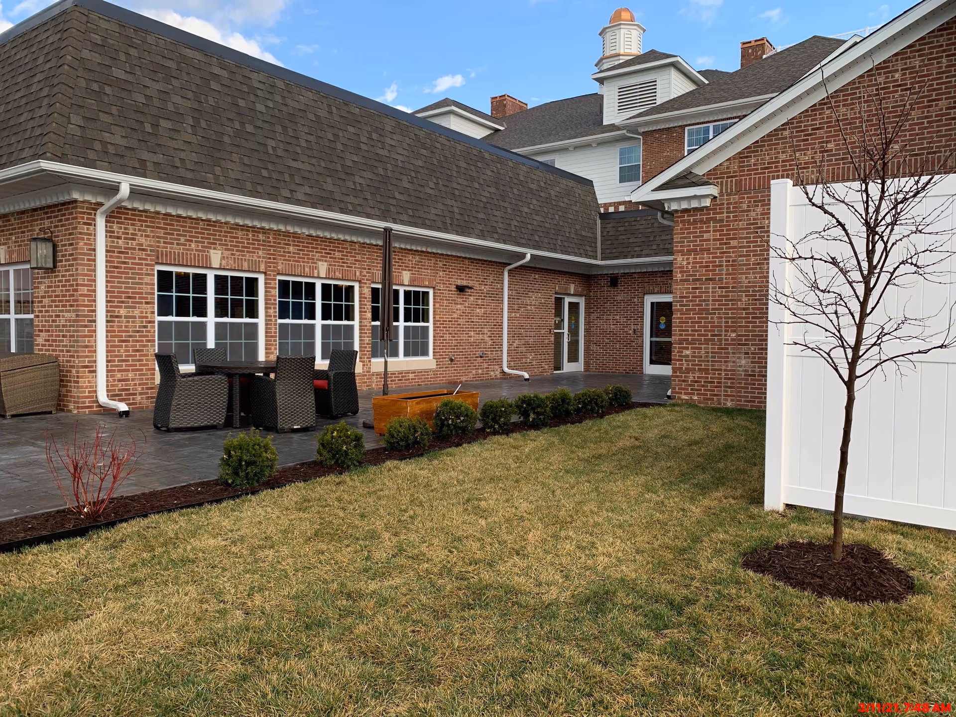 Courtyard patio with brick buildings, an outdoor dining table and chairs, small landscaped shrubs, and a white privacy fence.