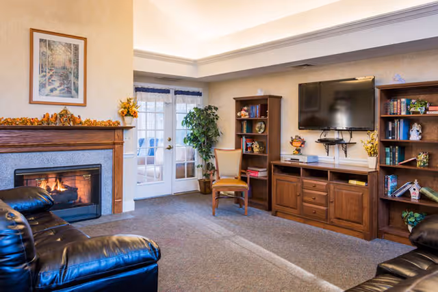 A cozy living room with leather sofas, a lit fireplace with autumn decorations on the mantle, a wooden entertainment center with a flat-screen TV mounted above it, two wooden bookshelves filled with books and decorative items, a wooden chair, and French doors leading to a sunroom or patio area.