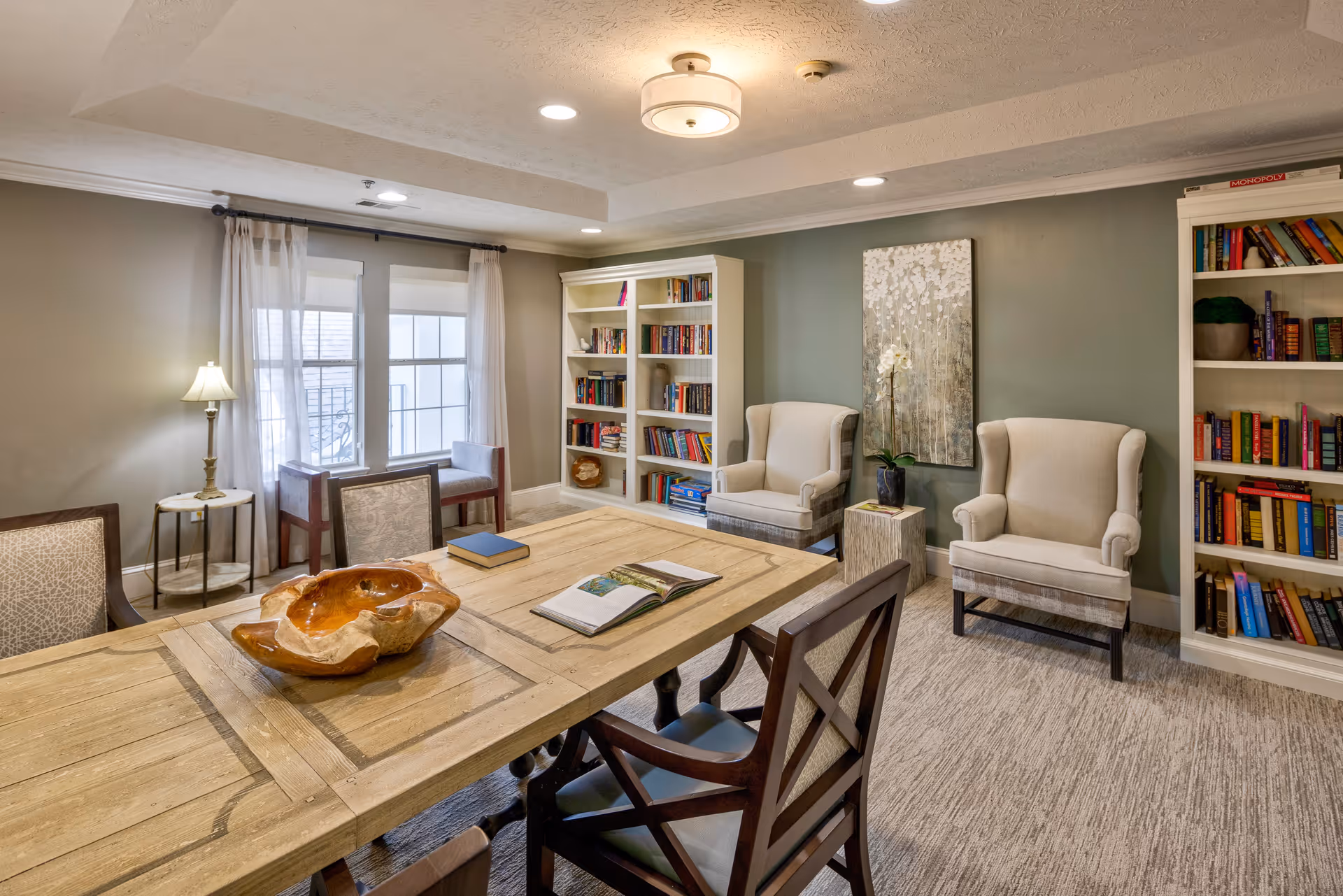 A cozy interior room with a large wooden table surrounded by chairs, two beige armchairs against a green wall, two white bookshelves filled with books, a window with sheer curtains, a floor lamp, and a piece of artwork hanging on the wall.
