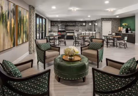 A modern common area with four armchairs arranged around a round green ottoman with a flower centerpiece. In the background, there are several small dining tables and chairs, a kitchenette with a coffee machine, and a patterned backsplash wall. Large abstract paintings hang on the left wall, and the room is well-lit with recessed ceiling lights.
