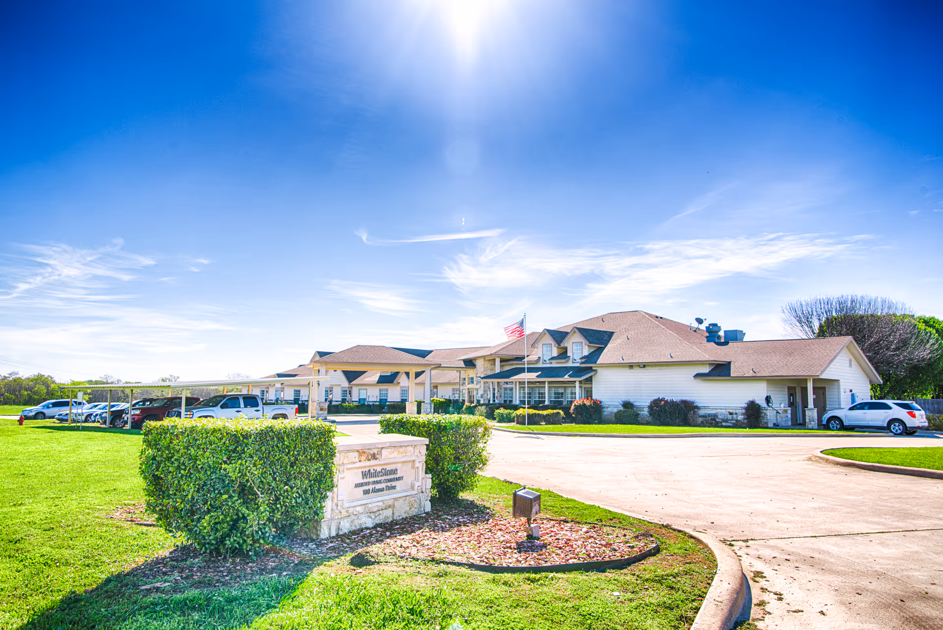 Exterior view of The Whitestone Assisted Living facility on a sunny day, showing a large building with a brown roof, an American flag, a parking area with several cars, and a green lawn with trimmed bushes and a stone sign displaying the facility's name and address.