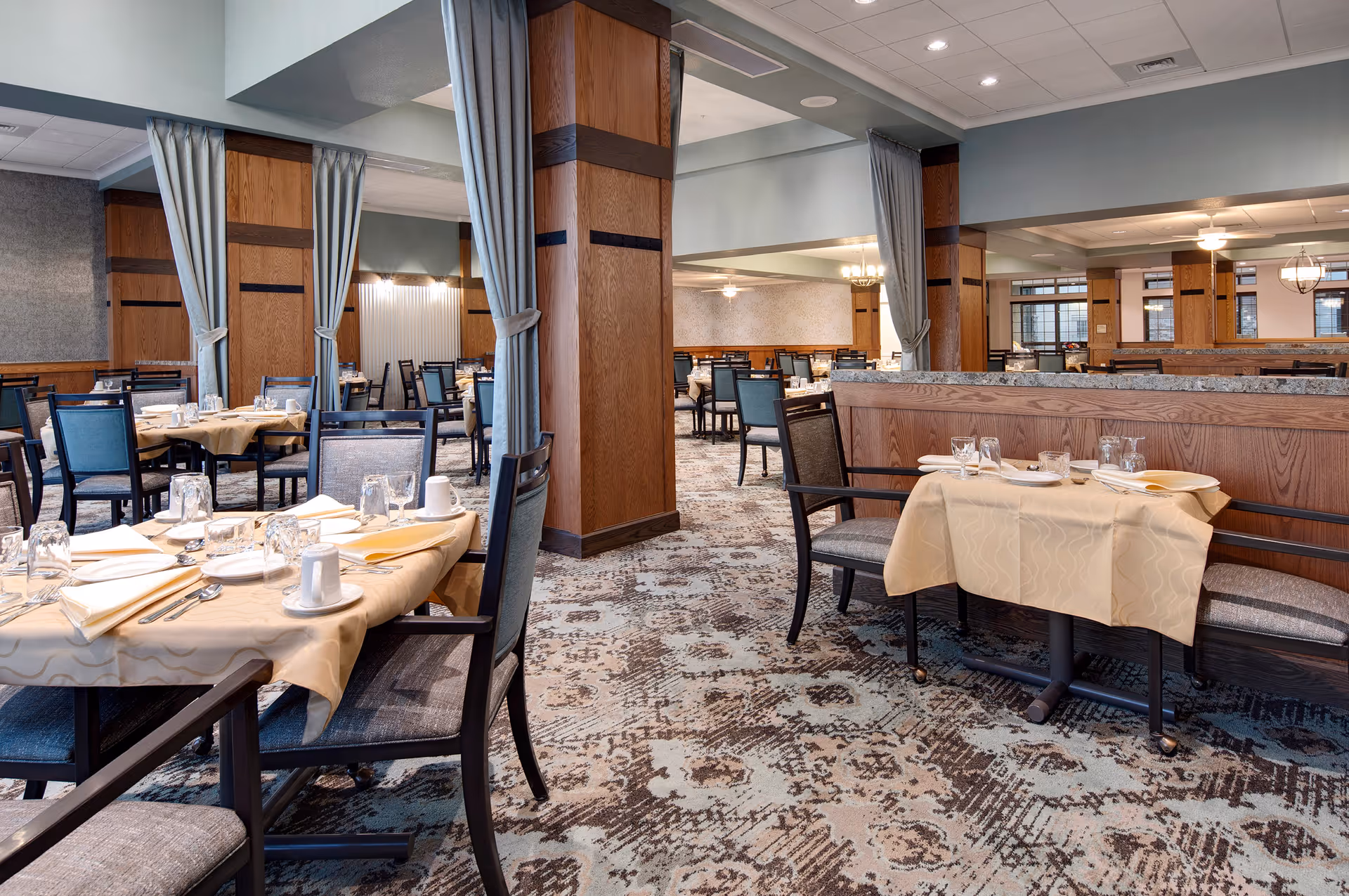 A spacious dining room in a senior living facility with multiple tables covered in beige tablecloths, set with plates, glasses, cups, and silverware. The room features wooden pillars with light blue curtains tied back, patterned carpet, and soft lighting from ceiling fixtures.