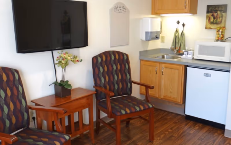 Two patterned armchairs and a small wooden table under a wall-mounted TV beside a kitchenette with a sink, microwave, and mini-fridge.