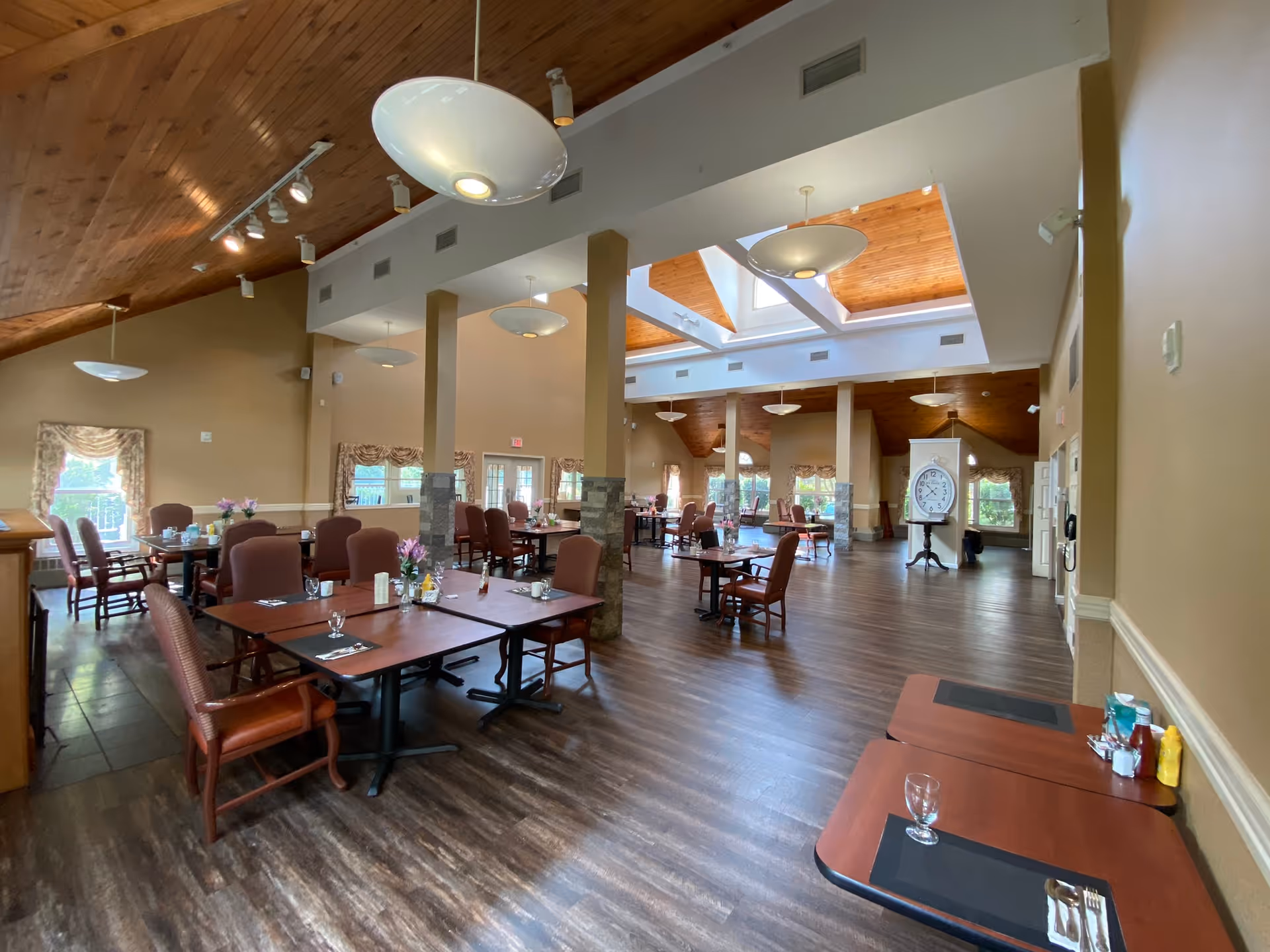 Spacious dining room with wooden floors and high ceilings featuring wooden paneling and skylights. Multiple tables with chairs are arranged throughout the room, some set with placemats, glasses, and condiments. Large windows with floral curtains allow natural light to enter the room. A large clock on a stand is visible in the background.