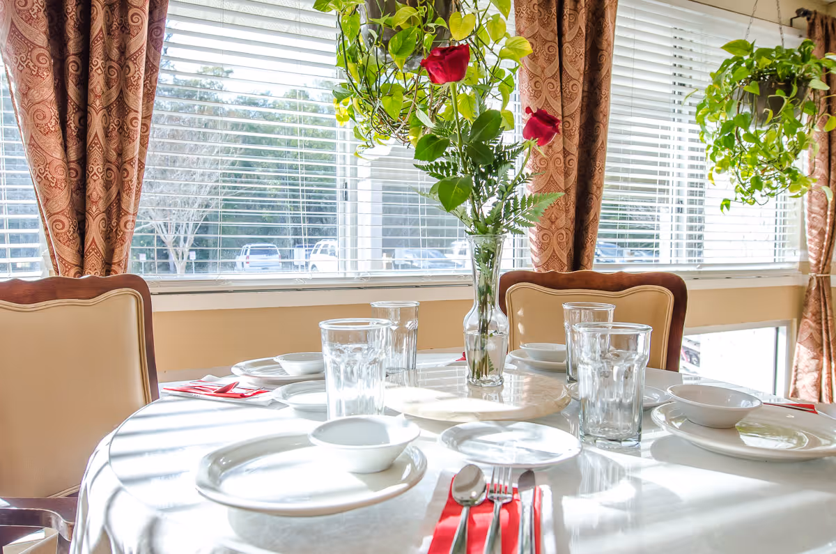 A round dining table set for six with white plates, bowls, glasses, and silverware on red napkins. A vase with red roses and green leaves is in the center of the table. The table is near large windows with white blinds and patterned curtains, with hanging green plants on either side.