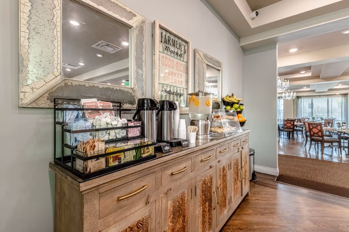 A wooden sideboard with a variety of coffee supplies, including creamers, sugar packets, stirrers, two coffee dispensers, a water dispenser with lemon slices, and a fruit basket. Above the sideboard are two rustic framed mirrors and a framed sign that reads 'Farmer's Market'. In the background, a dining area with tables and chairs is visible.