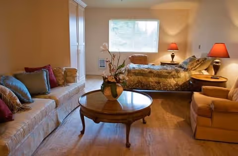 A cozy room with a bed covered in a patterned quilt, a wooden round coffee table with a flower vase, a beige sofa with colorful cushions, an armchair, two side tables with red lamps, and a large window letting in natural light.