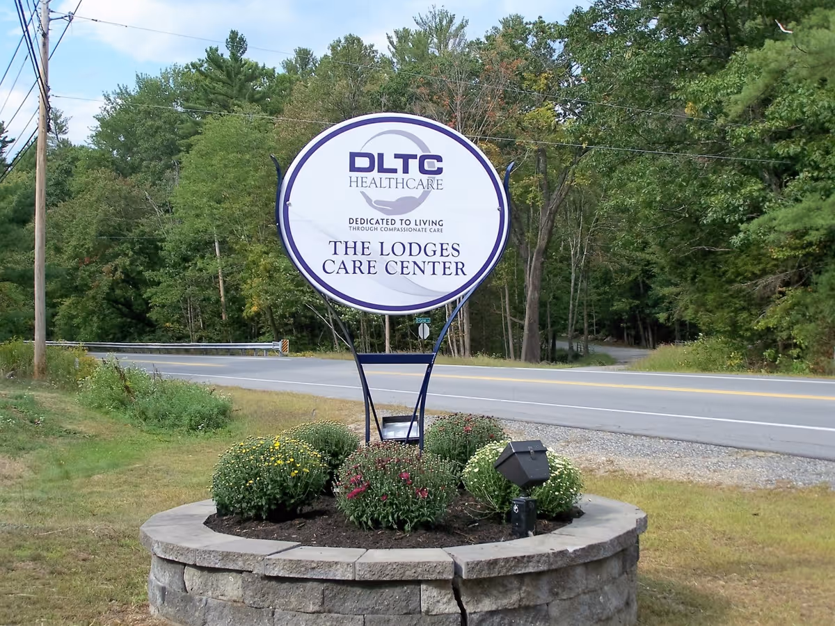 Circular roadside sign reading 'DLTC Healthcare The Lodges Care Center' in a stone planter with a road and trees behind.