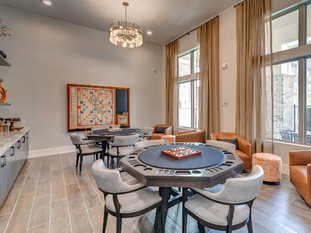 A bright and spacious game room with large windows covered by beige curtains. The room features two octagonal game tables with built-in cup holders and comfortable gray chairs around them. On the wall, there is a large Scrabble board and shelves with decorative items. The floor is covered with light wood tiles, and there are orange armchairs and small round ottomans near the windows.