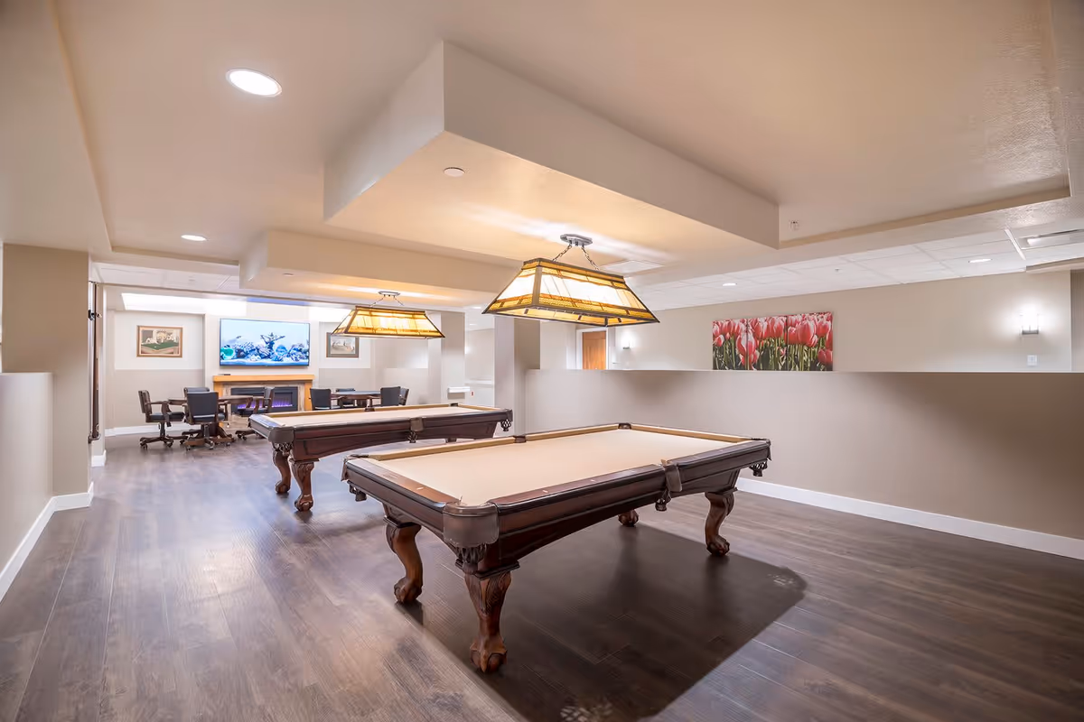 Well-lit game room with two pool tables, hanging lamps, and a seating area with a TV and fireplace.